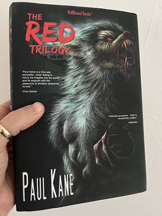 photograph of a hand with a gold thumb ring holding up a copy of the book The RED Trilogy by Paul Kane. A dark cover features a werewolf howling, with text in red