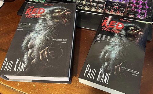 image showing a hardback and paperback copy of The Red Trilogy by Paul Kane, on a scratched wooden surface