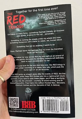 photograph of a man's hand holding up the rear cover of The RED trilogy by Paul Kane