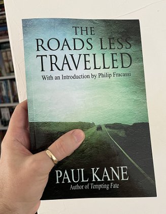 photograph of a man's hand holding a copy up of The Roads Less Travelled by Paul Kane, showing the front cover - a car disappearing into the distance down a long, straight road