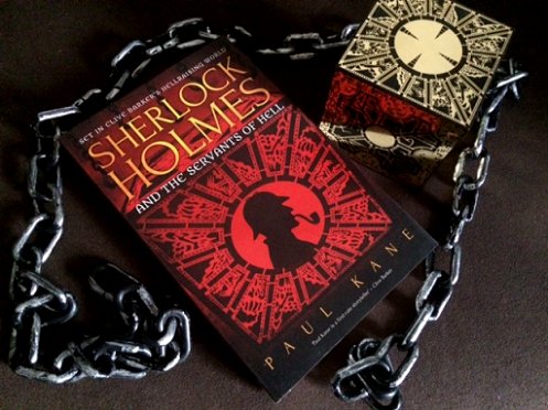 image showing a copy of Sherlock Holmes and the Servants of Hell by Paul Kane lying on a black surface. On its right is a Lemarchand configuration puzzle box, and both are wrapped in chain