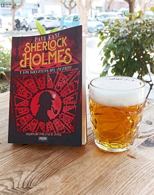 photograph of a standing copy of the Spanish edition of Sherlock Holmes and the Servants of Hell by Paul Kane, ona  wooden table beside a glass of beer