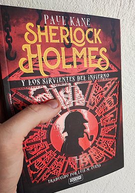 A man's hand holding a copy of the Spanish edition of Sherlock Holmes and the Servants of Hell by Paul Kane up against a cream wall