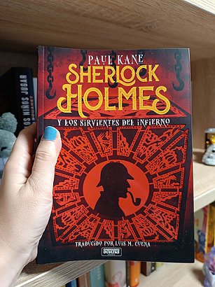 photograph of a hand with blue-painted nails holding up a copy of the Spanish edition of Sherlock Holmes and the Servants of Hell by Paul Kane, against a bookcase in the background