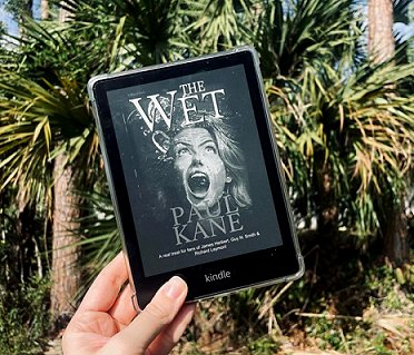 photograph of a hand holdinga  Kindle up against a background of a bush. Screen shows the cover of The Wet by Paul Kane