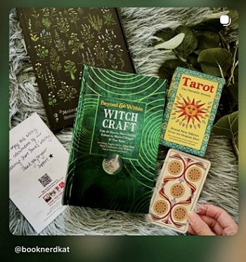 photograph showing a woman's hand holding a tarot card with the back of the card facing up, to the right of a copy of Beyond and Within Witchcraft, edited by Marie O'Regan and Paul Kane. In the background is a black and green journal, green leaves, a green fake fur rug and green leaves