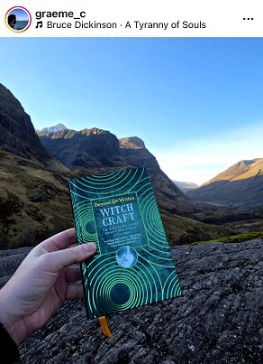 screenshot from @greaem_c. Picture shows a hand holding a copy of Beyond and Within Witchcraft, edited by Marie O'Regan and Paul Kane, up against a rocky hillside. The peaks of various hills can be seen in the distance