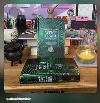 screenshot from @abookbumble. Photo shows a copy of Beyond and Within Witchcraft, edited by Marie O'Regan and Paul Kane, standing on another copy on a wooden surface. Surrounding the books are: a miniature black cauldron, a mug containing pens, a lit jar candle, and a laptop
