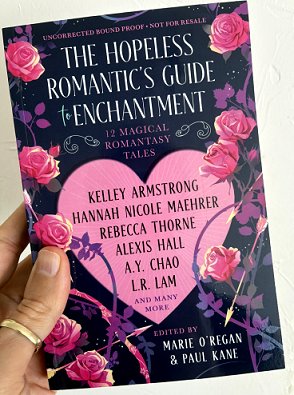 photograph of a man's hand holding a copy of The Hopeless Romantic's Guide to Enchantment, edited by Marie O'Regan and Paul Kane, up against a white surface