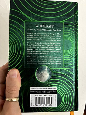 photograph of a man's hand holding up a copy of Beyond & Within: Witchcraft, edited by Marie O'Regan and Paul Kane, to show the back of the book